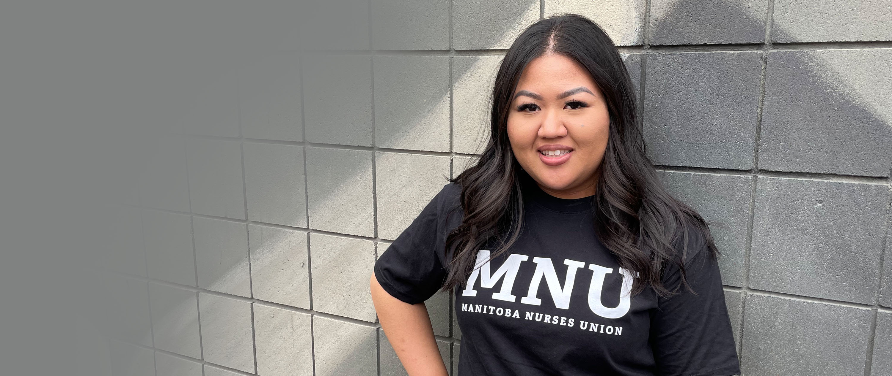 Woman standing against brick wall wearing a black t-shirt with the MNU Varsity Series logo